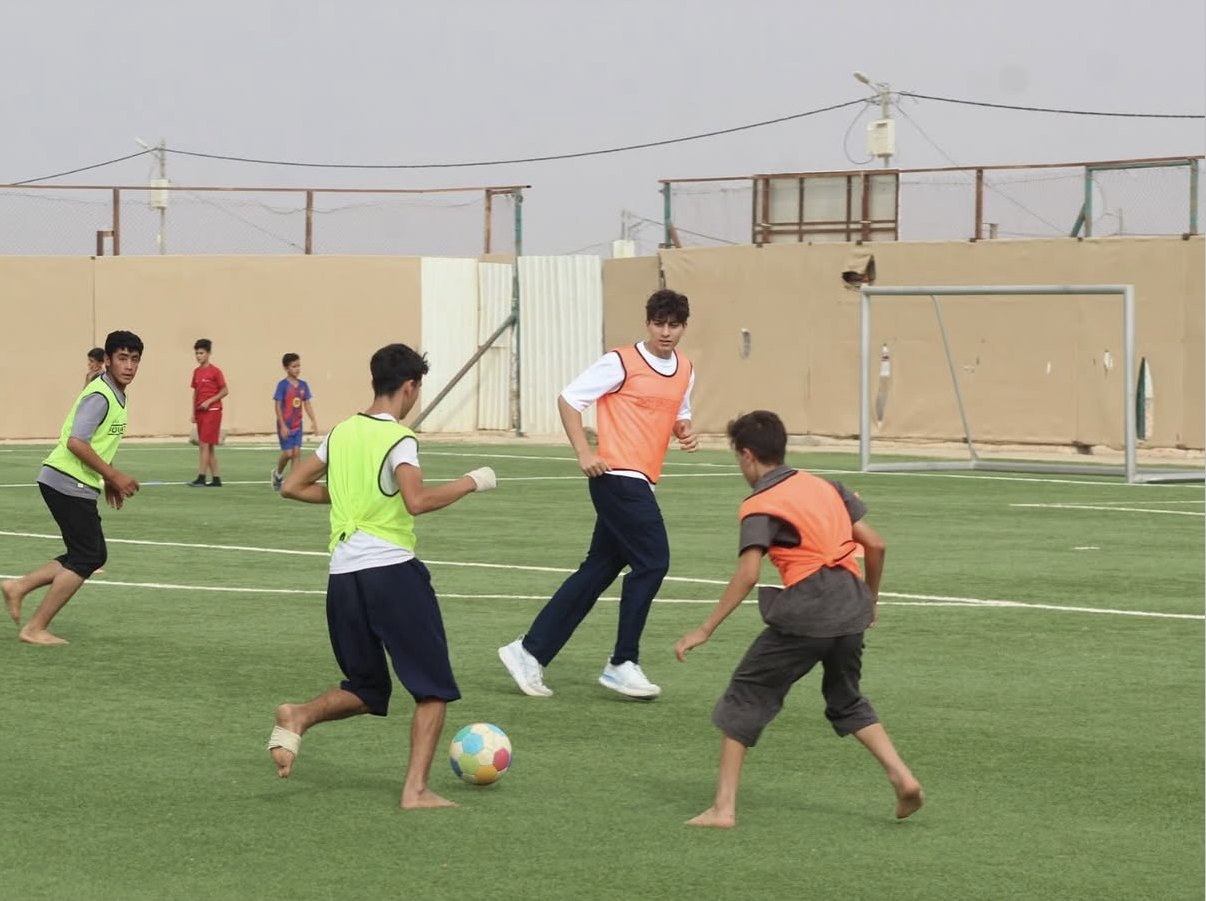Kids playing football on a turf pitch at Zaatari camp, several of them barefoot, with a Boots4Hope volunteer in the game